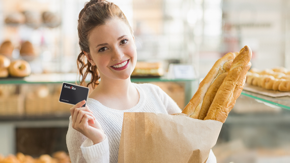 [Translate to English:] Frau in Bäckerei hat Tüte mit Baguette im Arm und hält bonVito-Kundenkarte hoch